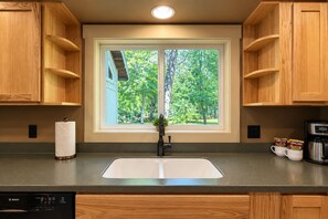 A kitchen with a double sink, wooden cabinets, a window with a view of trees, and a coffee maker. - Riverside Cabin-River, Hot Tub, WIFI, Dogs OK (Leavenworth)