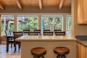 A kitchen with a large island, wooden chairs, and large windows with a view of trees.