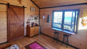L’intérieur d’une cabane est doté d’un plafond en bois, d’une grande porte coulissante, d’un coin cuisine, de deux tabourets, d’une fenêtre offrant une belle vue et d’un lit recouvert d’une couverture à motifs.