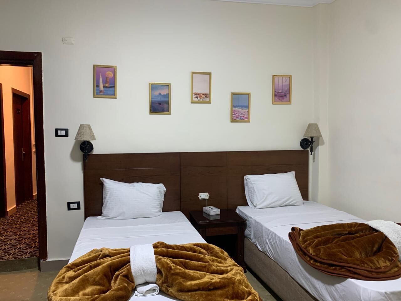 A hotel room with two single beds, a wooden headboard, a brown blanket on one bed, and four framed pictures on the wall.