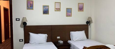 A hotel room with two single beds, a wooden headboard, a brown blanket on one bed, and four framed pictures on the wall.