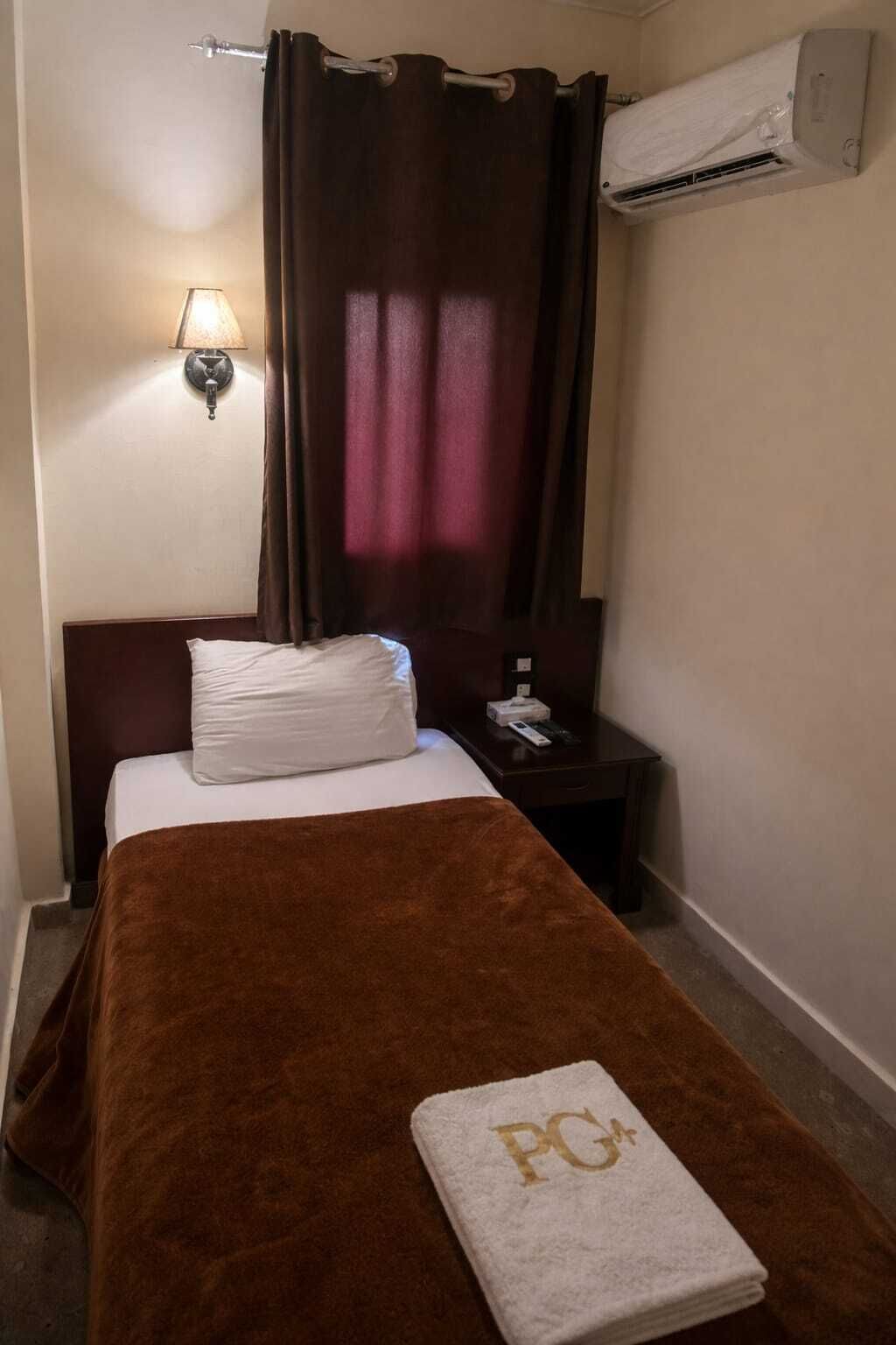 A hotel room with a bed, a brown blanket, a white pillow, a nightstand with a lamp, and an air conditioning unit.