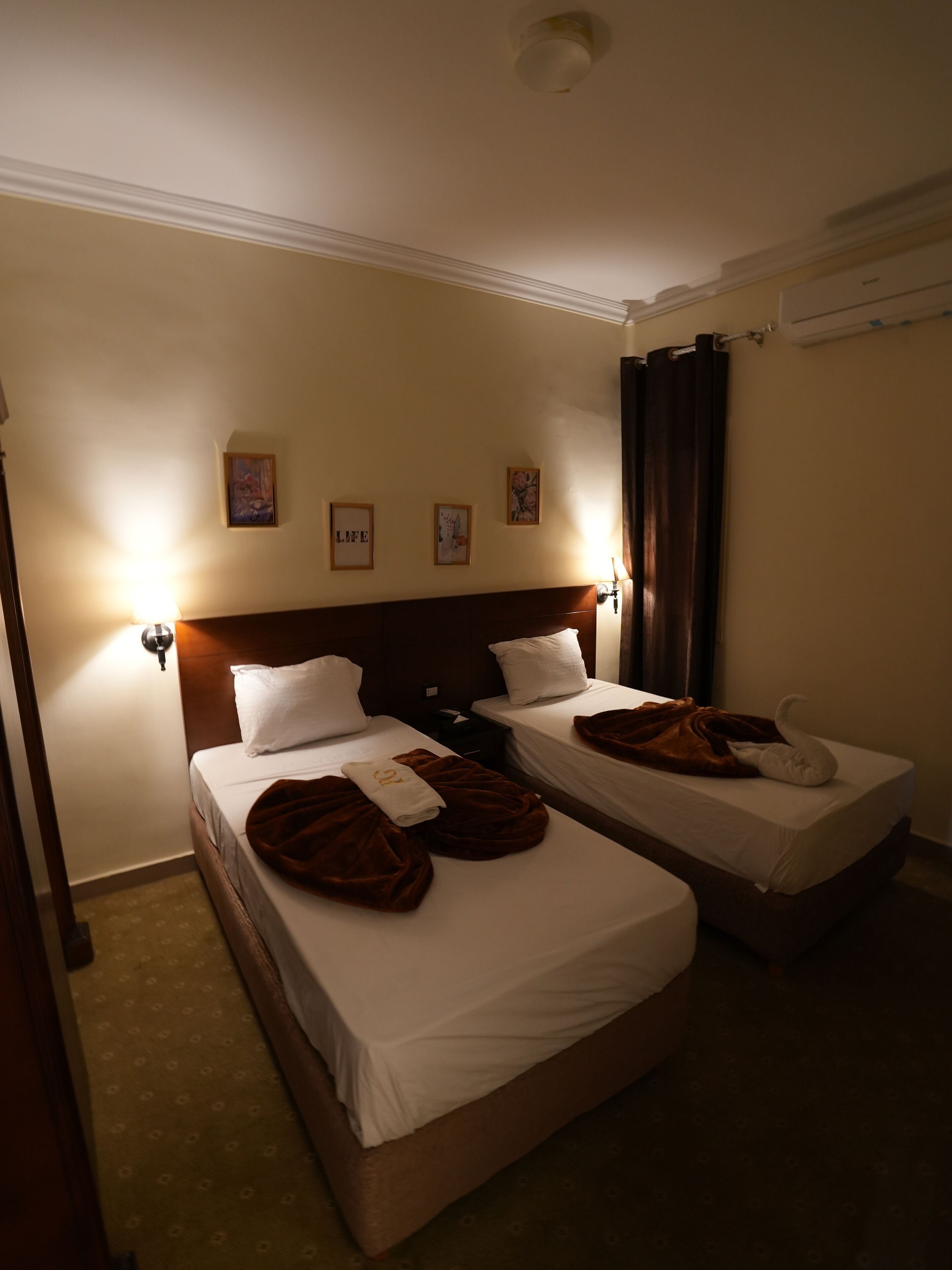 A hotel room with two single beds, a wooden headboard, a wall-mounted air conditioner, and framed artwork on the wall.