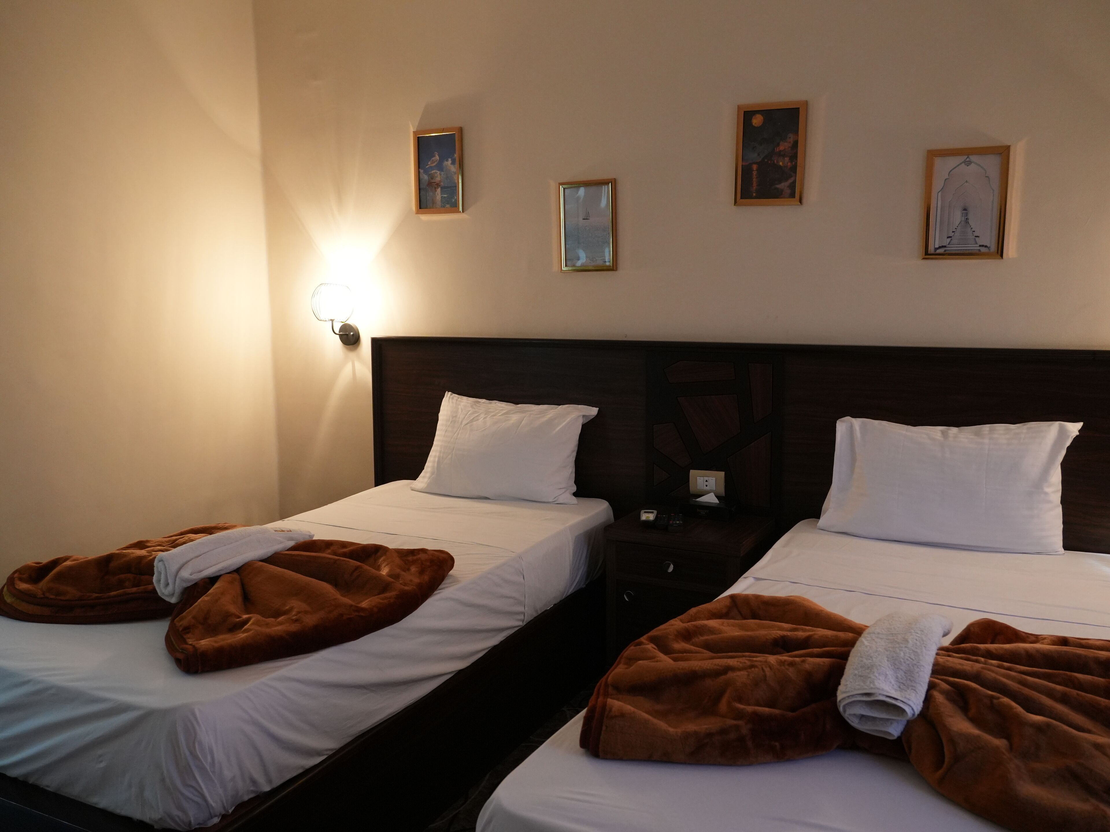 Une chambre d’hôtel avec deux lits, chacun étant équipé de draps blancs et de couvertures marron. Des appliques murales et des tableaux encadrés sont fixés au mur au-dessus des lits.