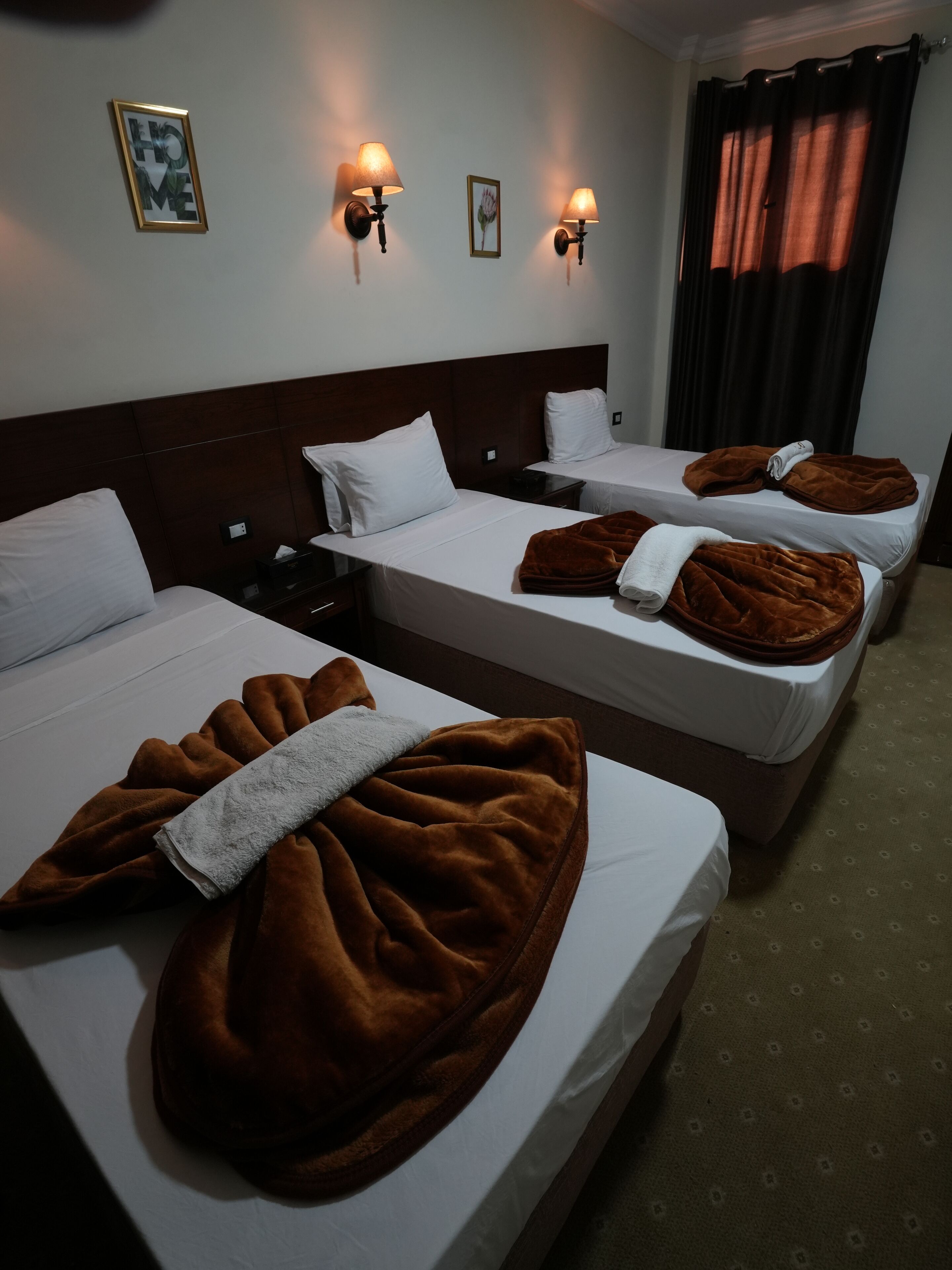 A hotel room with three beds, each with white linens and brown blankets. There are wall-mounted lamps and a framed picture on the wall.