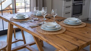 Uma mesa de jantar de madeira posta com pratos, tigelas, talheres e copos de vinho, com uma cozinha ao fundo.
