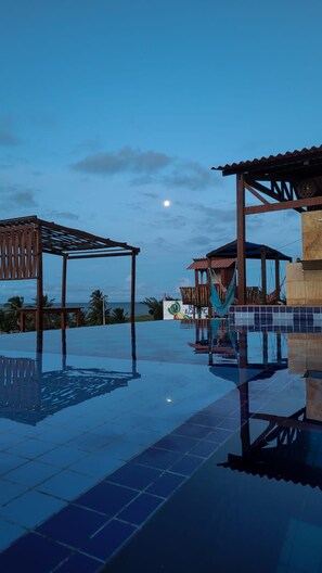 An infinity pool with wooden pergolas and a hammock, reflecting the sky and surroundings.