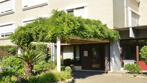 En byggnads entré med en överbyggd veranda, en liten trädgård med växter och en bänk.