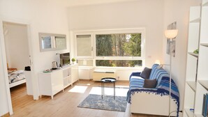 Ett kompakt bostadsutrymme med ett litet kök, en soffa, ett matbord och ett stort fönster med utsikt över omgivningen.