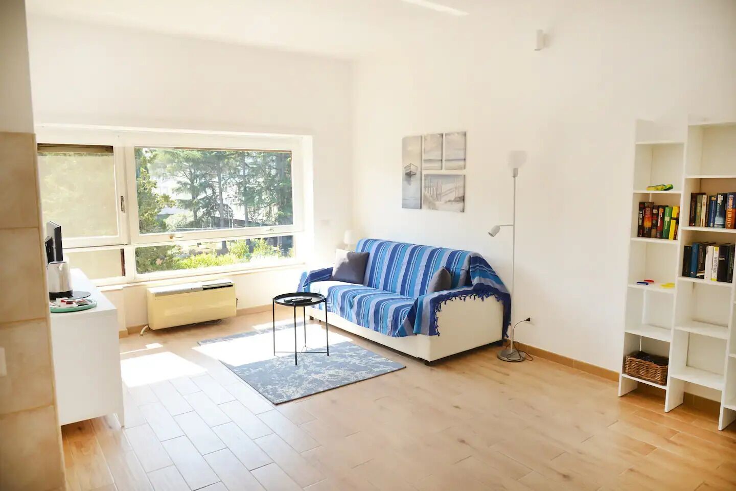 Ein modernes Wohnzimmer mit einem weißen Sofa, einem Couchtisch, einem Bücherregal und einem großen Fenster mit Blick auf Bäume.