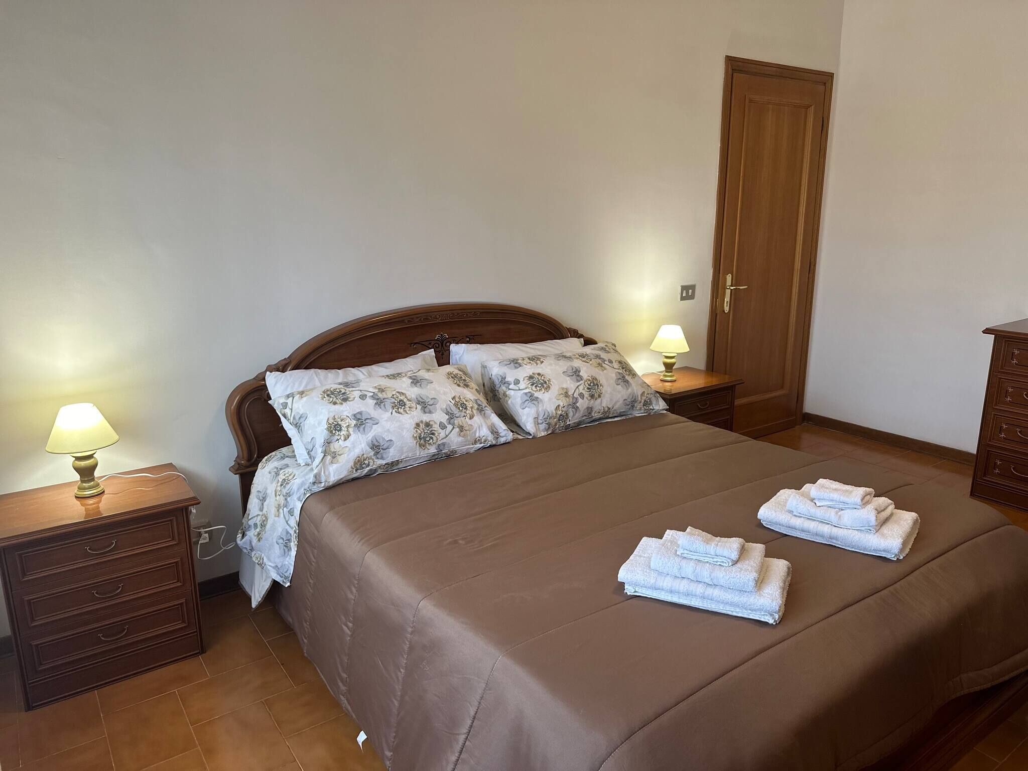 Ein ordentlich bezogenes Bett mit floralem Bettzeug, zwei Nachttischlampen und eine Reihe Handtücher auf dem Bett.