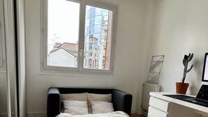 Une chambre à coucher avec un lit, un bureau, une plante en pot et une fenêtre donnant sur des immeubles.