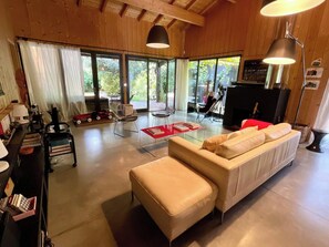 Ein großzügiges Wohnzimmer mit einer breiten Couch, einem Couchtisch und einem Fernseher. Vor den Fenstern hängen Vorhänge, und eine Lampe ist sichtbar.