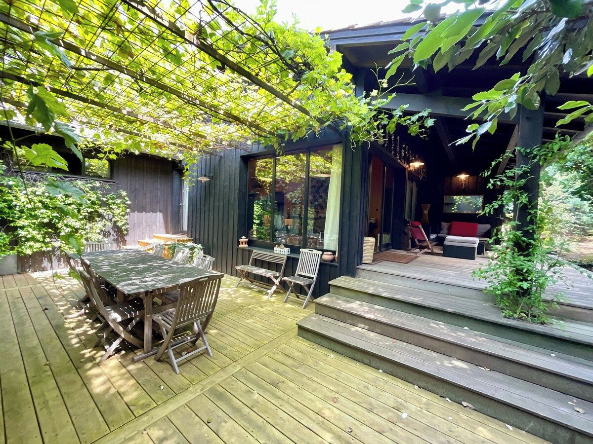 Eine Holzterrasse mit Tisch und Stühlen, einem überdachten Sitzbereich und einer Treppe, die zu einem Gebäude führt.
