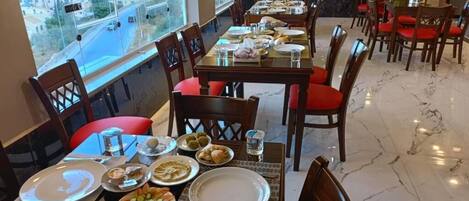Un comedor con varias mesas dispuestas para comidas, ventanas amplias que ofrecen vistas de un paisaje urbano, y una pantalla de televisión mostrando un noticiero.