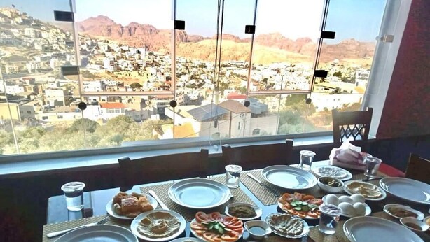 A dining table set with plates, bowls, and cutlery, overlooking a scenic view of a town and mountains. - Petra Crystal hotel (Wadi Musa)
