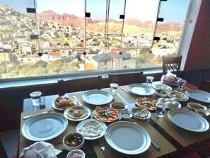 A dining table set with plates, bowls, and cutlery, overlooking a scenic view of a town and mountains. - Petra Crystal hotel (Wadi Musa)