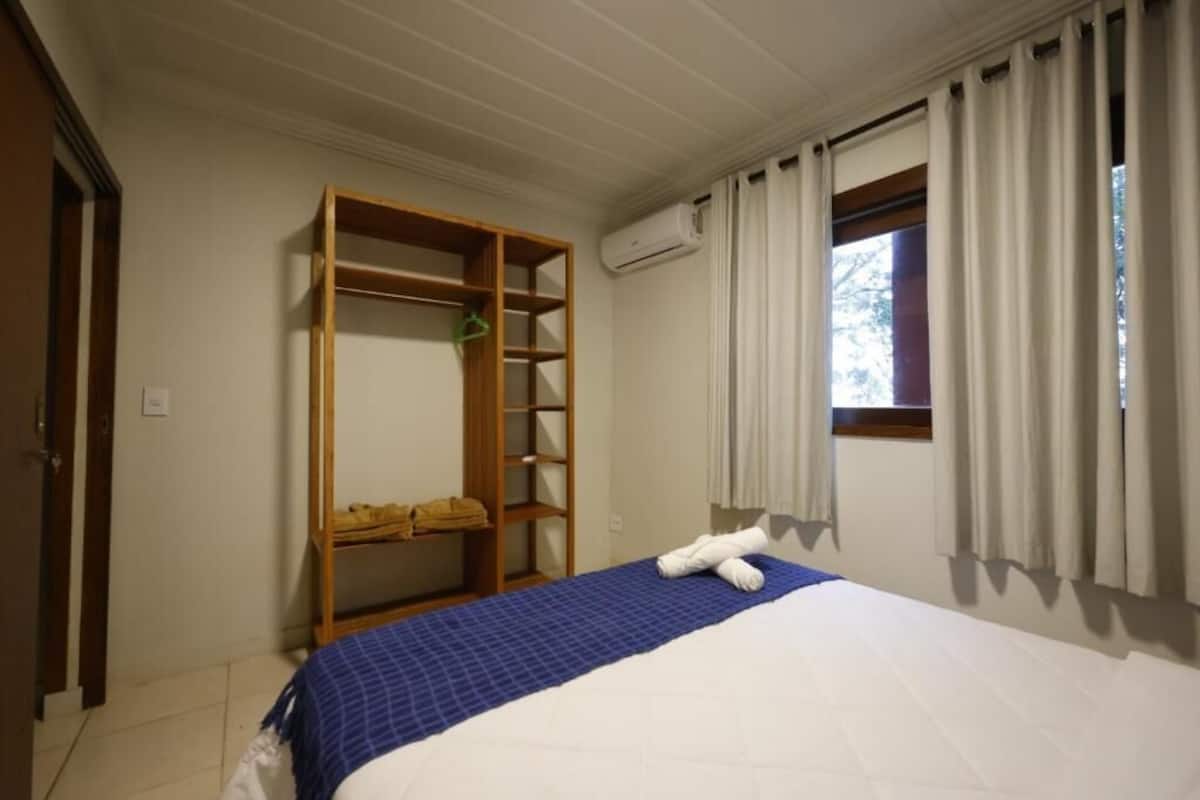 A hotel room with a bed, a wooden wardrobe, a window with curtains, and an air conditioning unit.