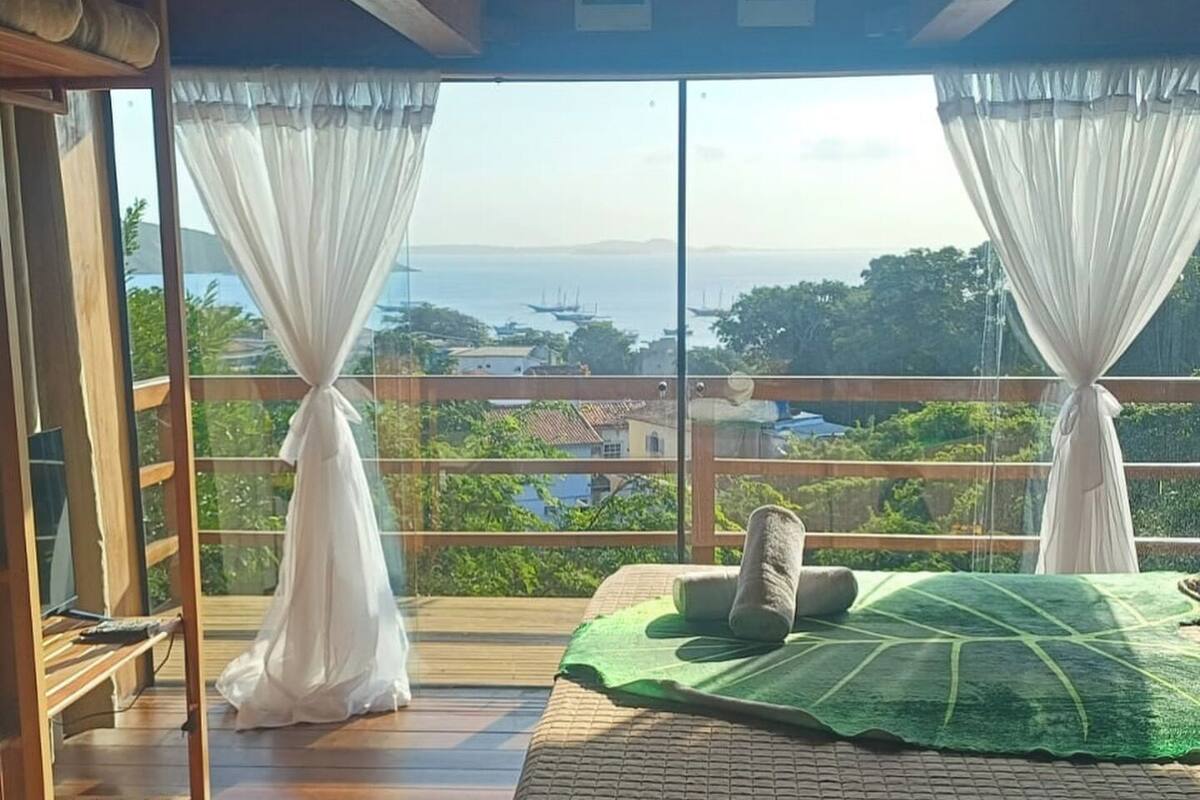 A bedroom with a wooden floor, a bed with a green towel, and a view of the sea and boats.