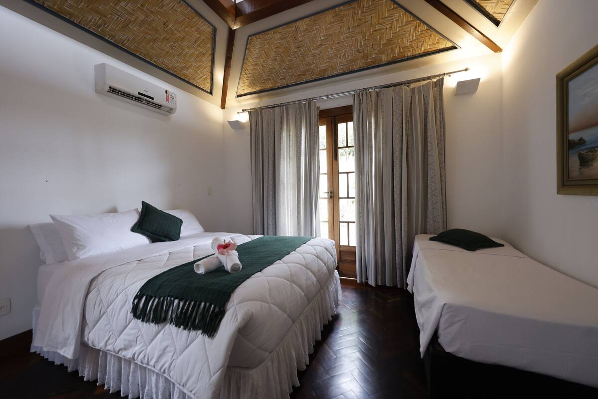 A bedroom with a large bed, two smaller beds, a window with curtains, a painting on the wall, and an air conditioning unit.