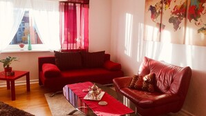 Una sala de estar con un sofá rojo, un sillón rojo, una mesa de centro con una bandeja, una mesita con una planta y una ventana con cortinas de gasa.