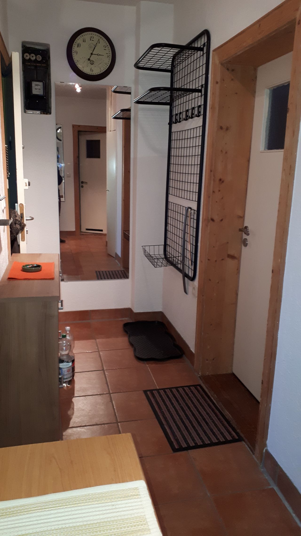 Un pasillo con puerta de madera, estante de metal negro, un reloj y suelo de baldosas.