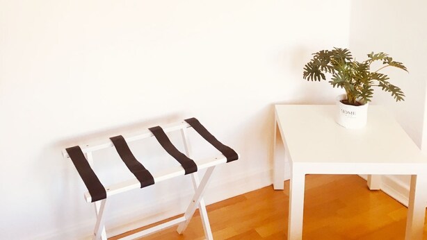 Una habitación con pared blanca, ventana con vista al mar, una mesita blanca, un banco blanco y negro, y una planta en maceta.