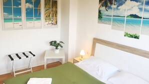 Habitación de hotel con una cama, una mesita pequeña con una planta, una mesita de noche con una lámpara y una obra de arte en la pared que representa escenas tropicales.