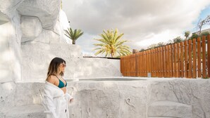 A person walking on a stone pathway beside a pool with a wooden fence and palm trees in the background.