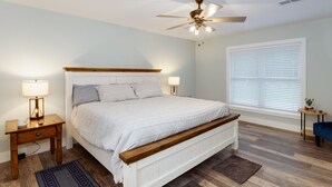 A bedroom with a wooden bed, two bedside tables with lamps, a ceiling fan, and a large window with blinds.