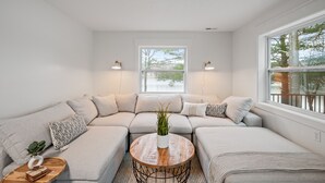 Un salon moderne avec un canapé d’angle, une table basse ronde en bois et une vue sur les arbres par une grande fenêtre.