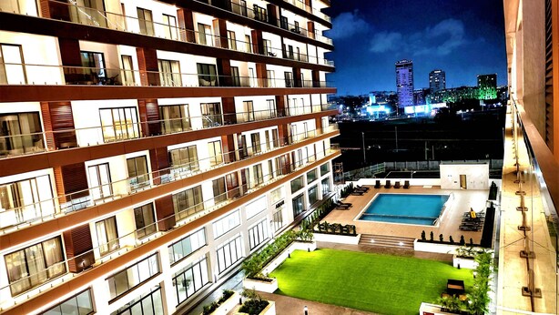 A multi-story hotel with a rooftop pool, a well-maintained lawn, and a cityscape view in the background.