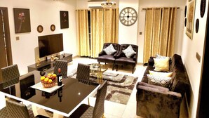 A modern living room with a dining area, featuring a flat-screen TV, a chandelier, and a large wall clock.