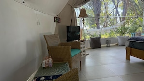 A cozy room with a wicker chair, a small table with magazines, a TV, and a bed with a view of trees through a window. - Amaj Eco Lodge (Sámara)
