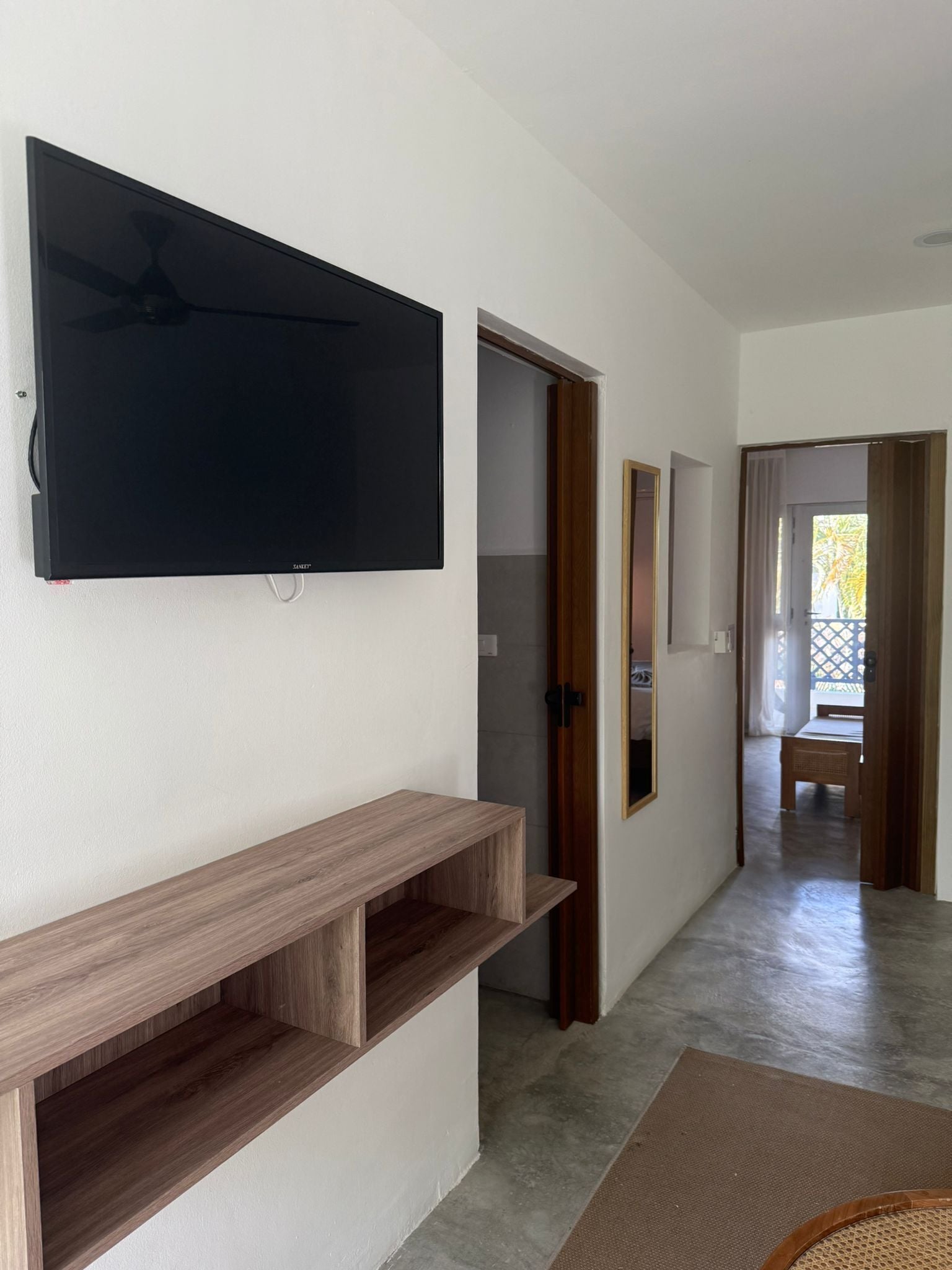 A hallway with a flat-screen TV mounted on the wall, wooden shelves, and a view into a room with a bed and a window.