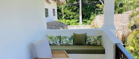 Un balcón con mobiliario de mimbre, una mesita redonda y vistas a vegetación exuberante y un edificio blanco.