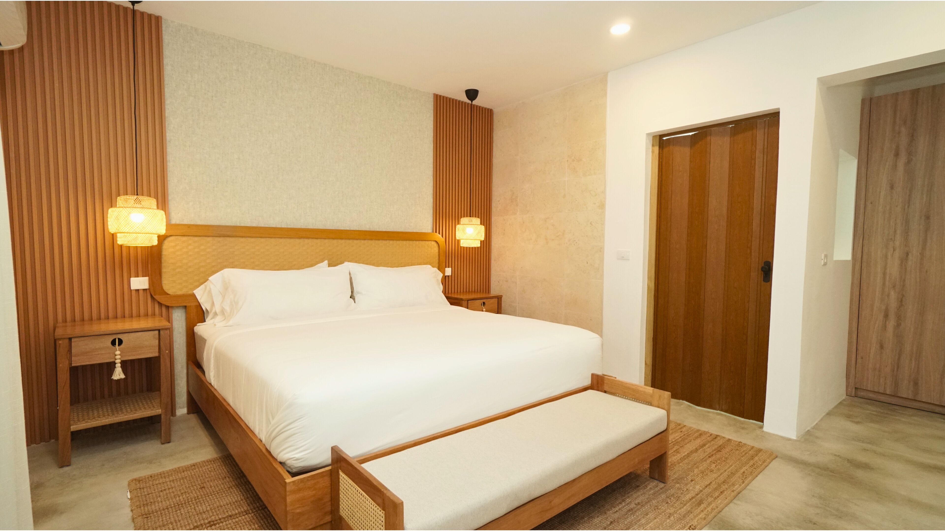 A modern hotel room with a wooden bed, two bedside tables with lamps, a wooden wardrobe, and a small bedside drawer.