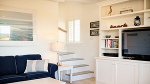 A living room with a blue sofa, a white coffee table, a TV on a white cabinet, and a staircase with a view to the outside. - Relaxing 3 bedroom cottage located in beautiful Prince Edward Island, Canada (Kensington)