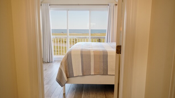 A bedroom with a large bed, a view of the beach, and a balcony with white curtains. - Relaxing 3 bedroom cottage located in beautiful Prince Edward Island, Canada (Kensington)