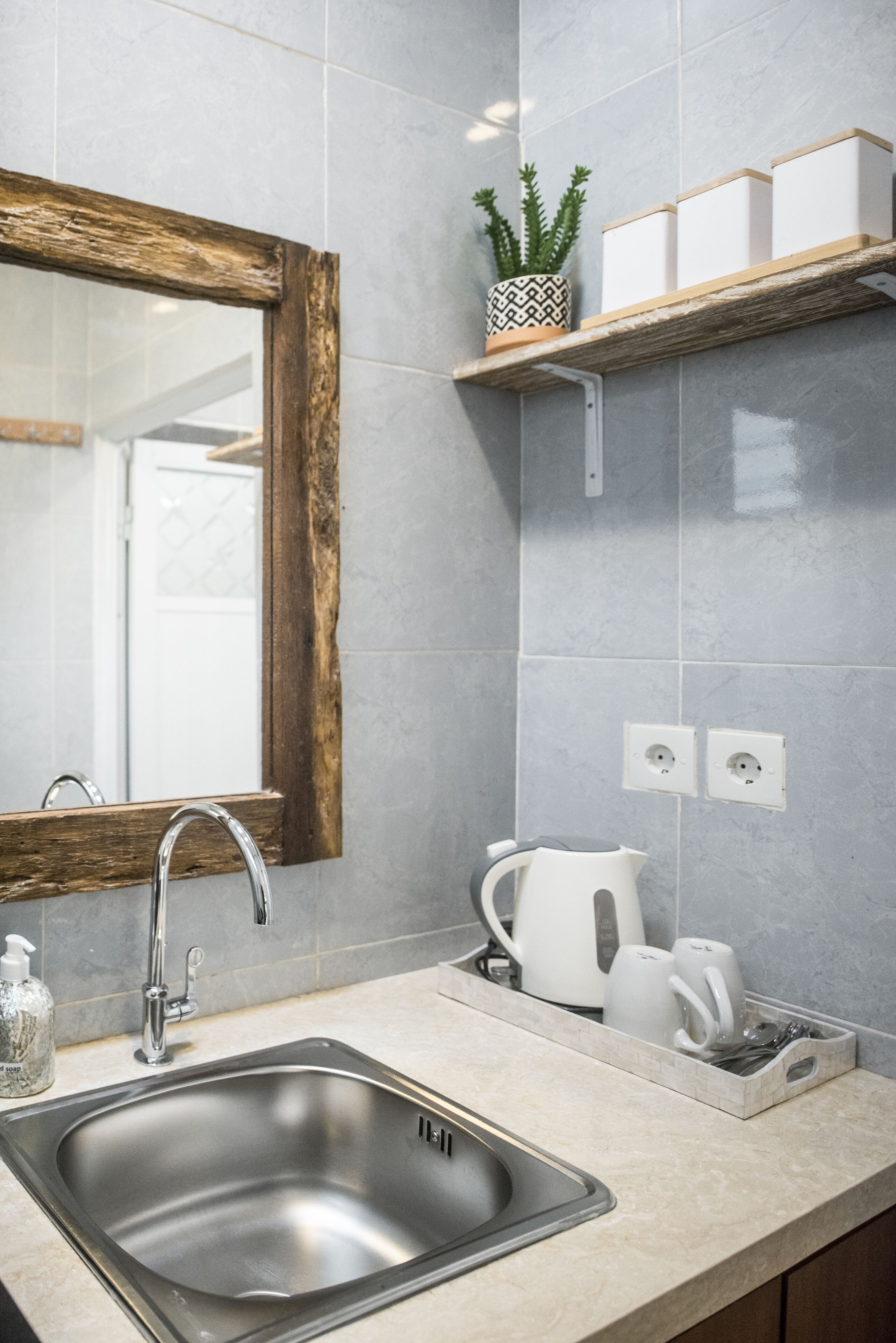 A modern bathroom with a large mirror, a stainless steel sink, a white kettle, and a potted plant on a shelf.