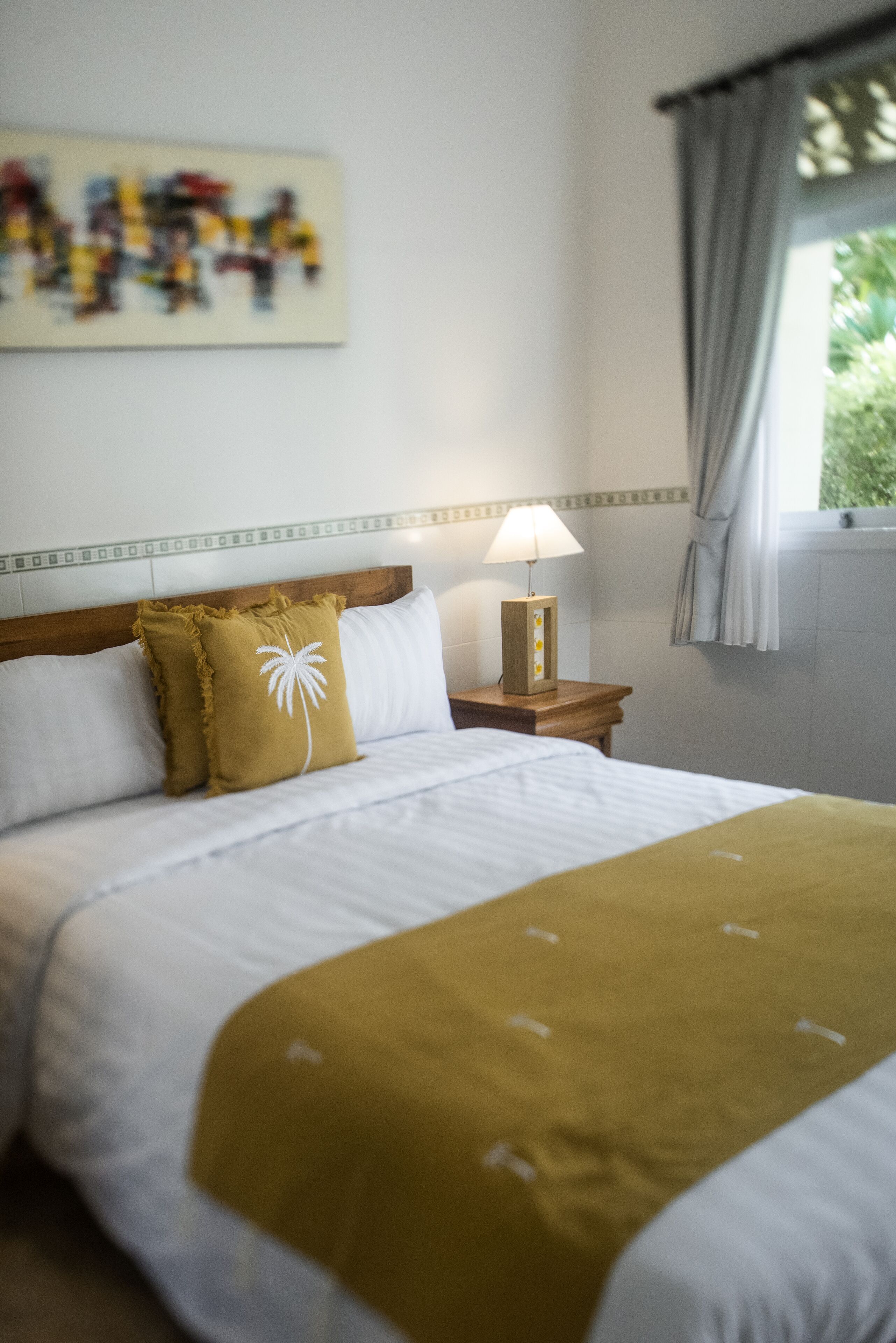 Una cama bien hecha con una colcha blanca y mostaza, una mesita de noche de madera con una lámpara, y un cuadro en la pared.