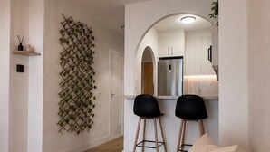 A modern living room with a sofa, a hanging light fixture, and a kitchen area visible through an archway.