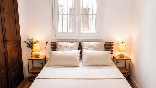 A bedroom with a large bed, two bedside lamps, a window with bars, and a wooden wardrobe.