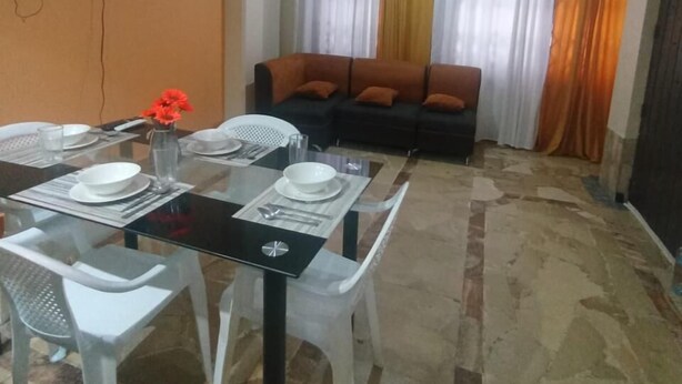 Un comedor con mesa de vidrio y sillas blancas, sofá con cojines anaranjados y televisor montado en la pared.