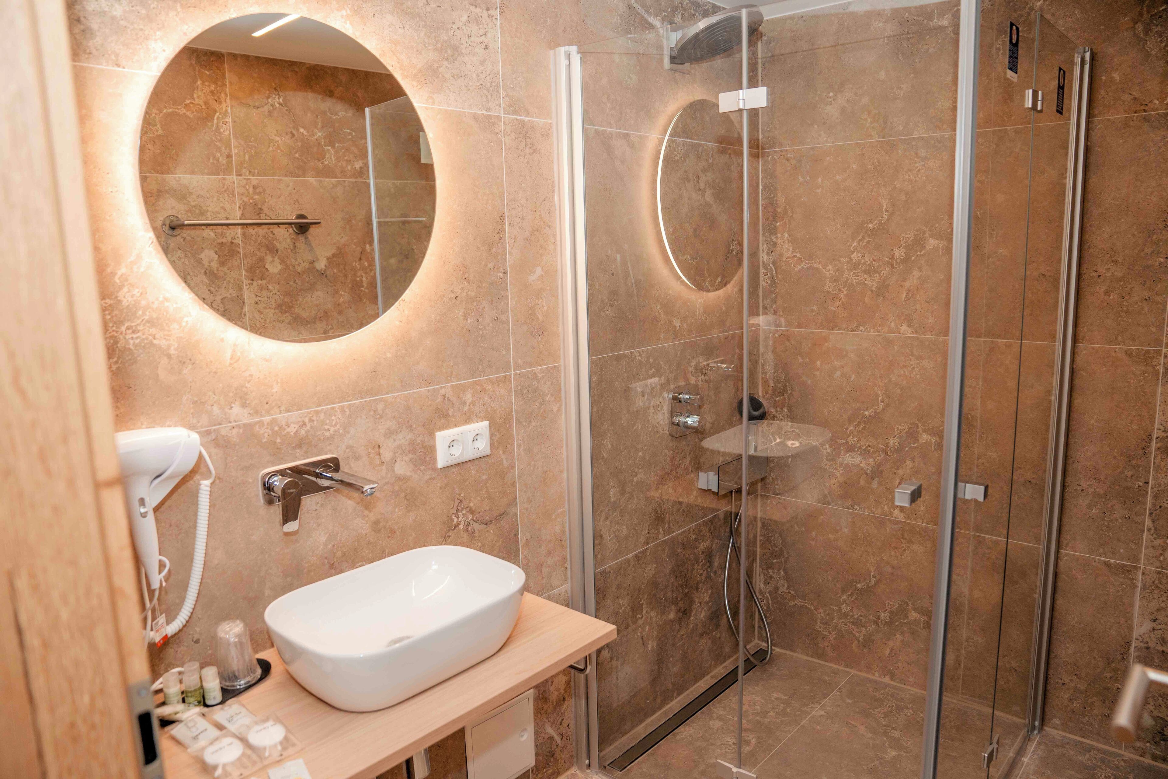 A modern bathroom with a large oval mirror, a white sink, and a glass-enclosed shower area.