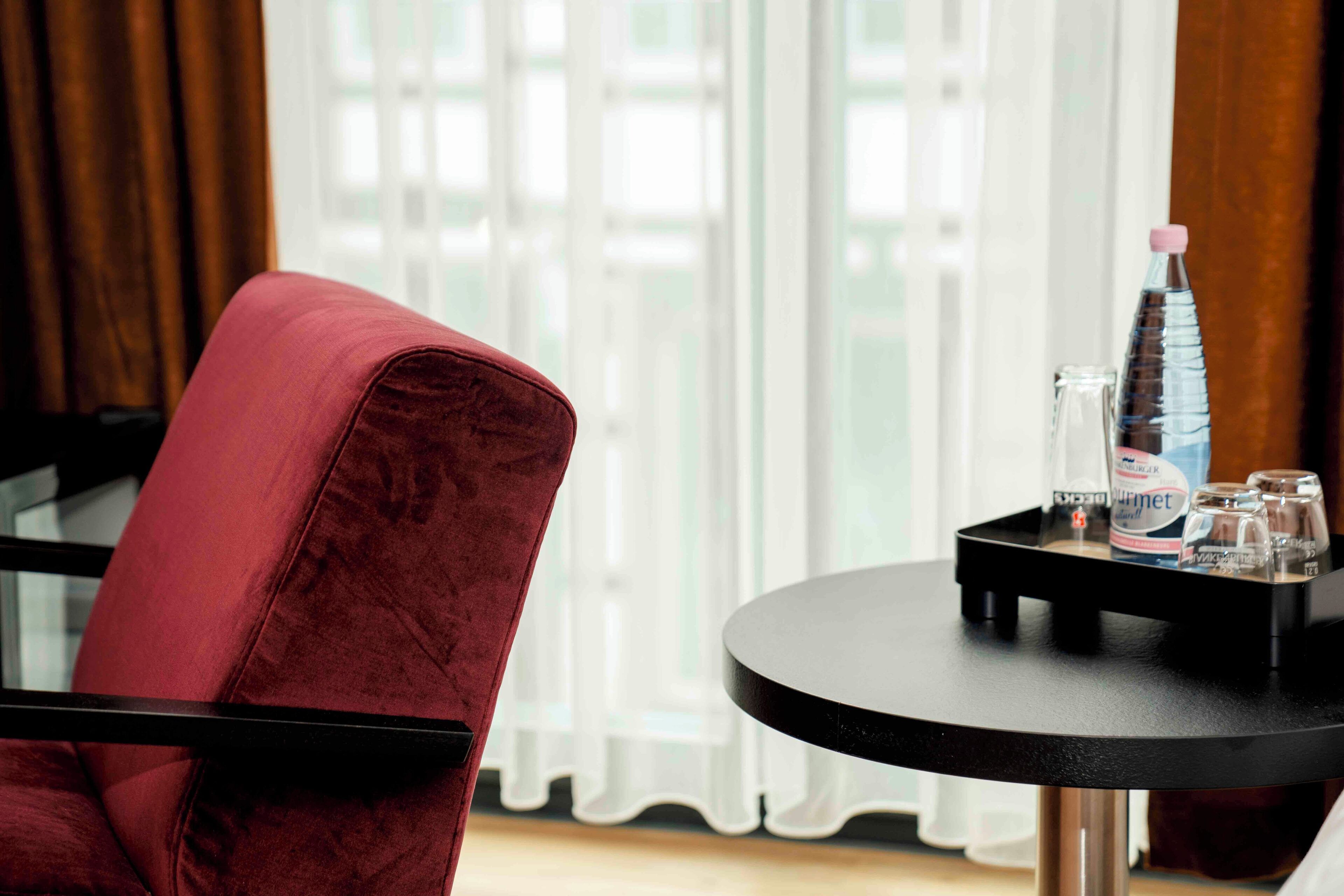 A room with a red velvet chair, a round black table with a bottle, glasses, and a water bottle.