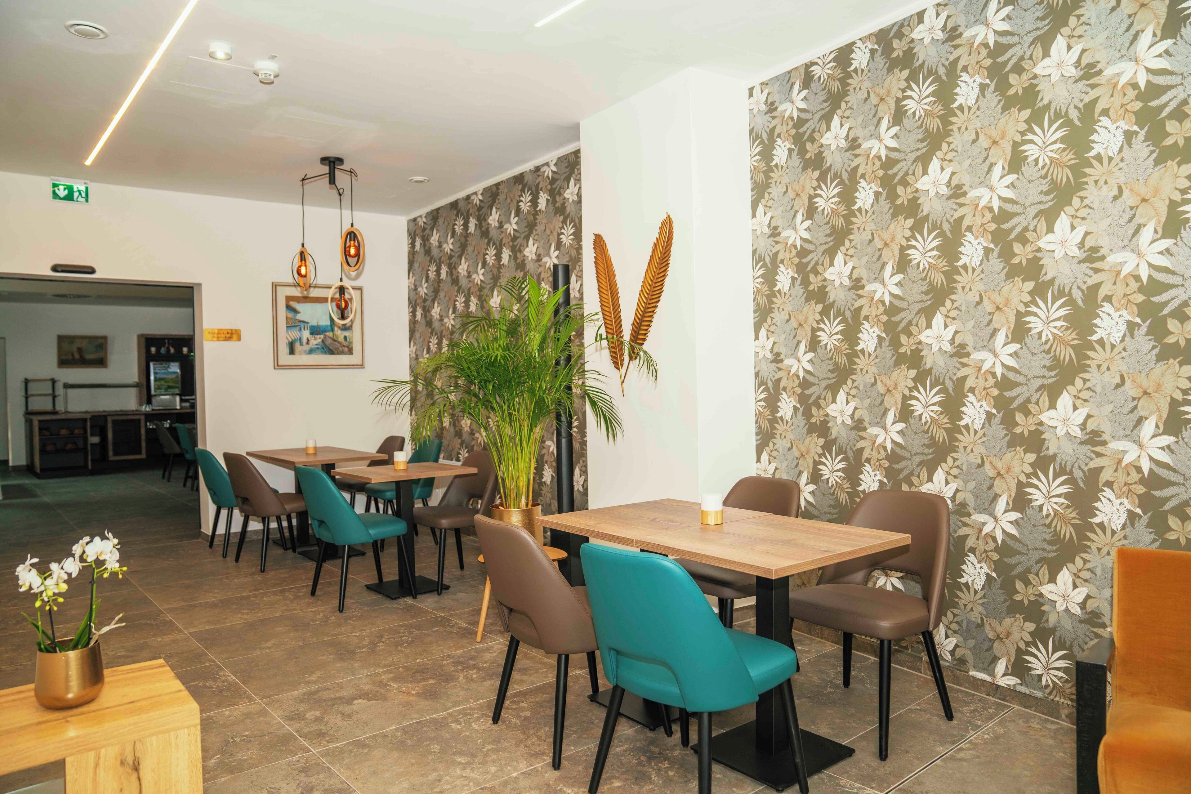 A hotel lobby with multiple dining tables and chairs, a potted plant, and a wall with a floral pattern.