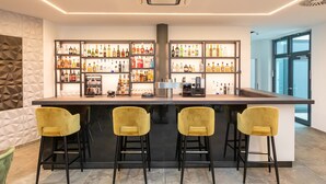Een moderne bar met een centraal pilaster, meerdere flessenrekken vol drank en drie gele barkrukken.