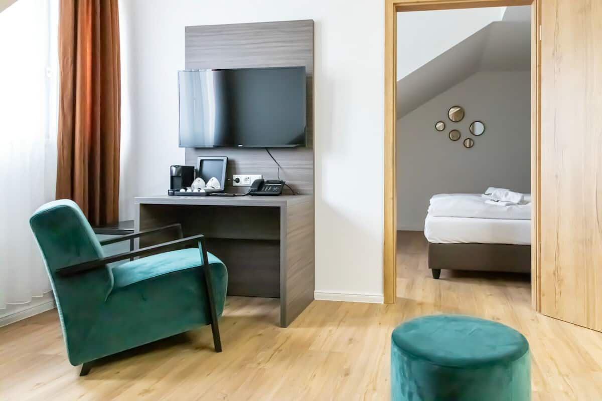 A modern hotel room with a teal armchair, a desk with a flat-screen TV, a bed, and a wooden wardrobe.