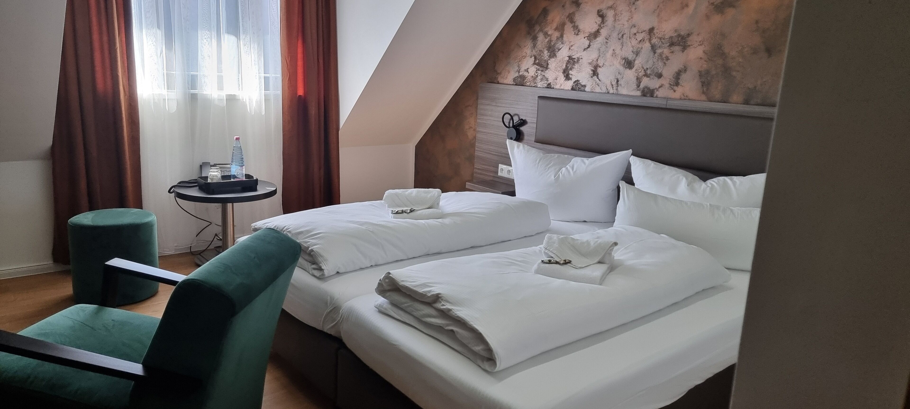 Una habitación de hotel con dos camas, una mesita redonda con una botella y un teléfono, una silla color turquesa y una ventana con cortinas.